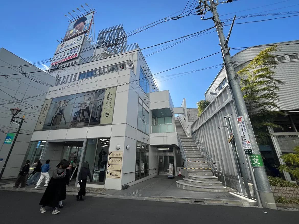 都会の街角にあるビルと階段、歩行者と店舗の看板、青空と都市の日常風景