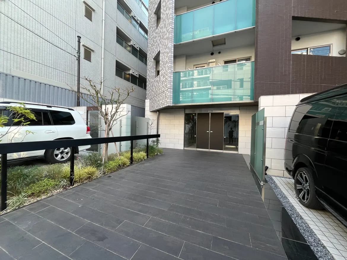 現代的な外観のマンションエントランス、植栽付きのアプローチ、黒い車が停車中