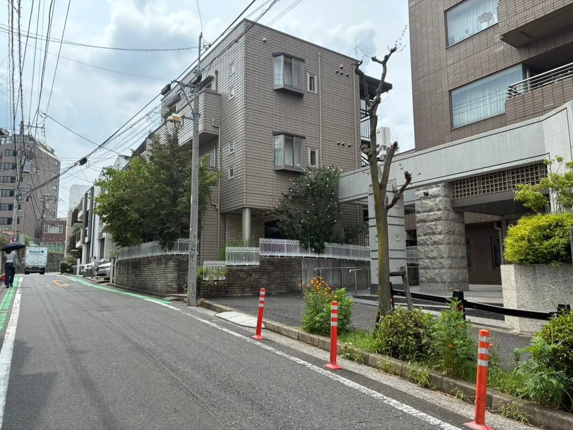 - 住宅街の静かな通り、周囲には高層ビルと緑の木々、歩行者が雨傘をさしている様子