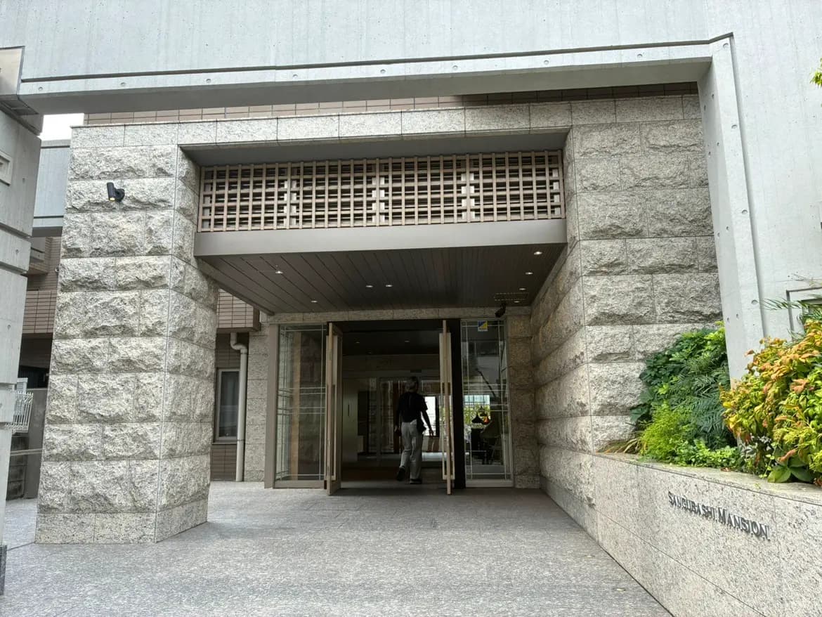 - モダンな外観のマンションエントランス、石造りの柱とガラスの自動ドア、周囲には緑の植物が配置されている景観