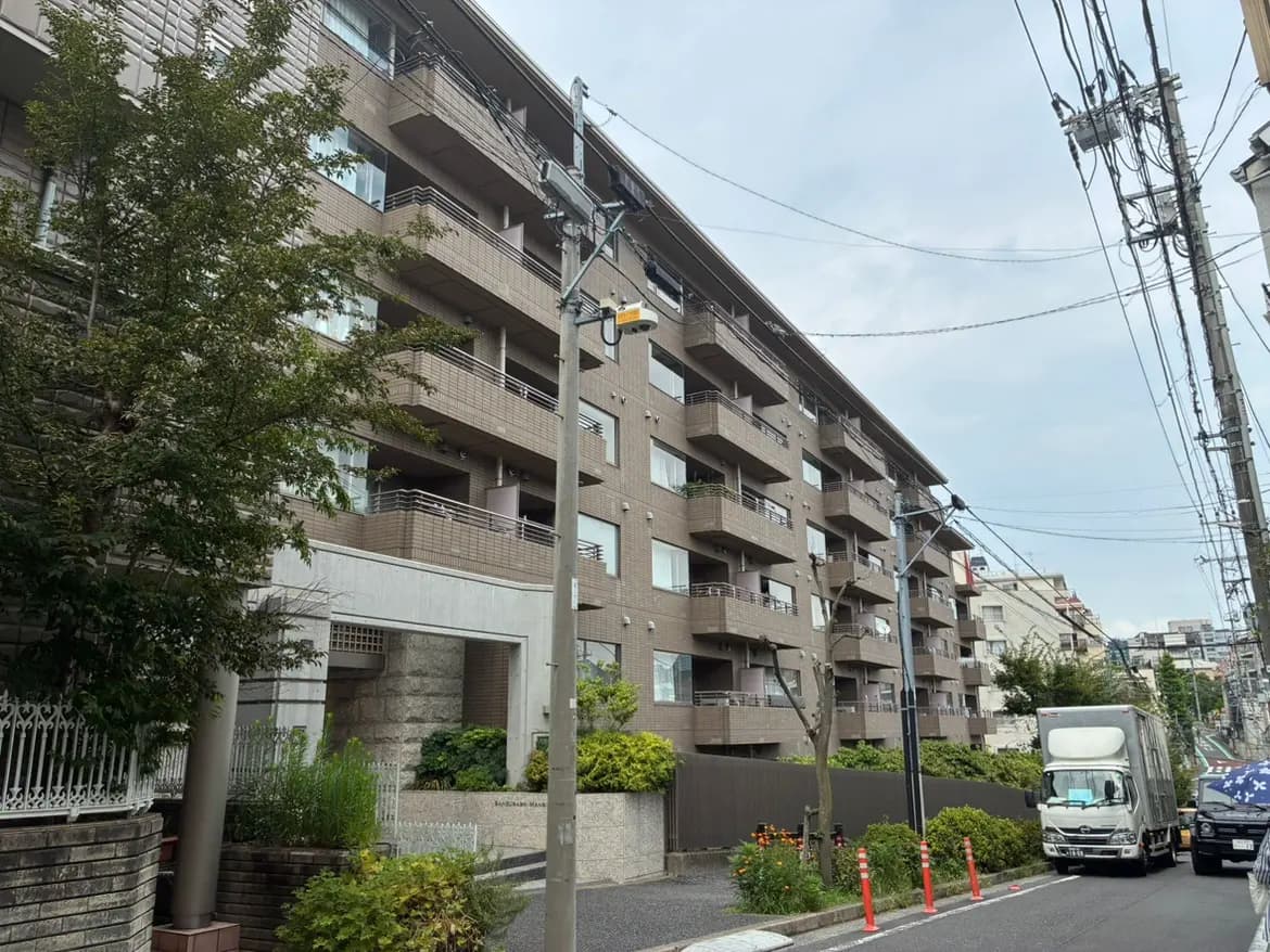 - 自然に囲まれた現代的なマンションの外観、バルコニーと大きな窓が特徴、周囲には緑豊かな植栽と道路が見える景観