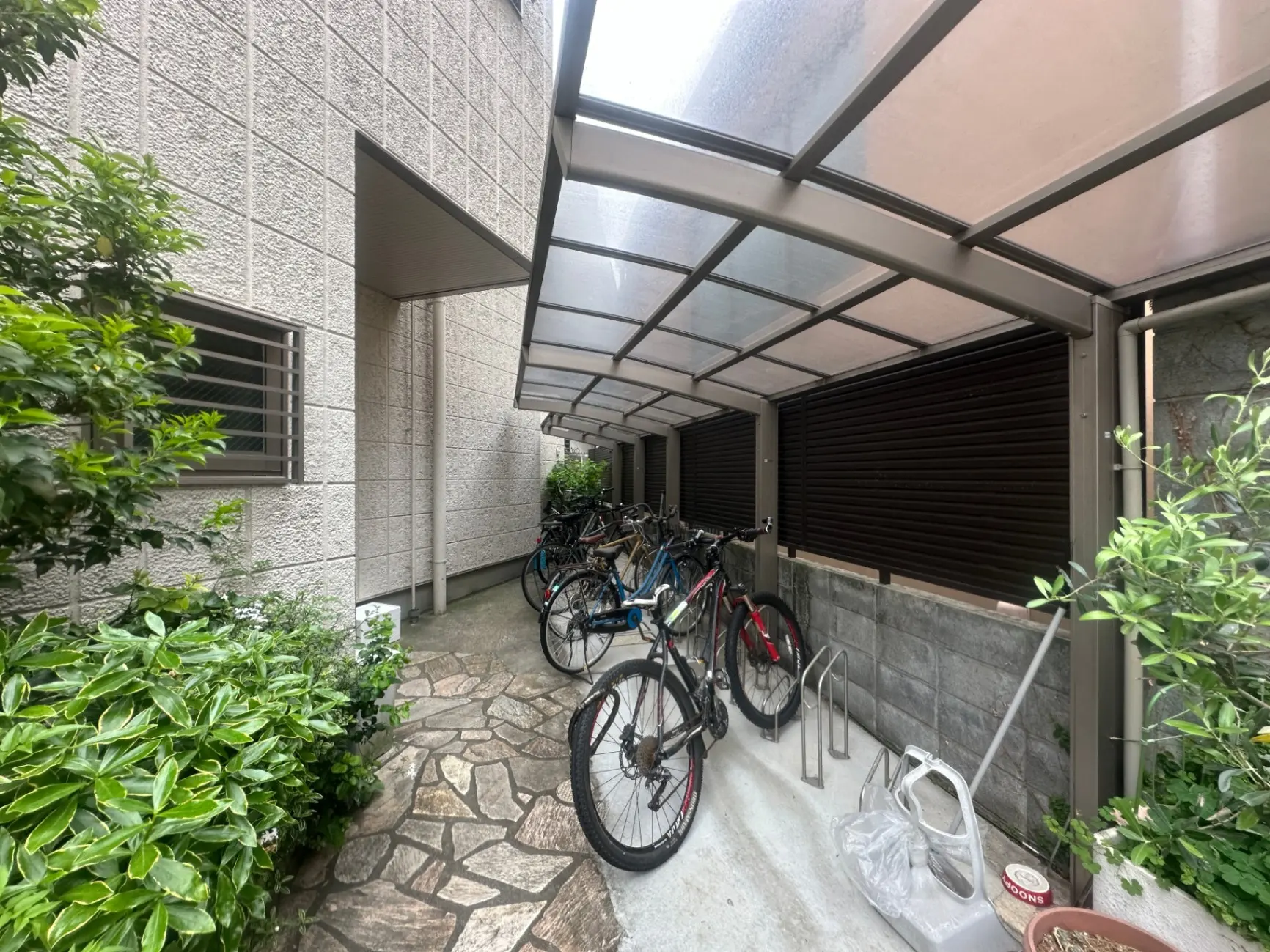 自転車置き場として利用されている屋根付きの小道、石畳の床と緑の植栽がある外観