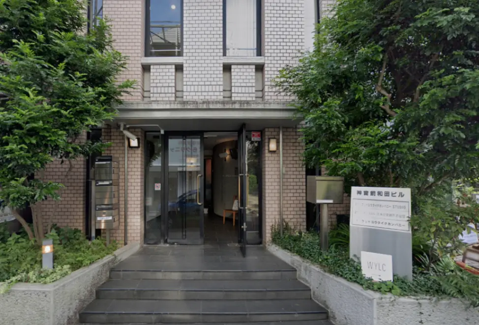 緑に囲まれた外観のオフィスビル、ガラスのエントリードア、会社名が書かれた看板のあるエントランス