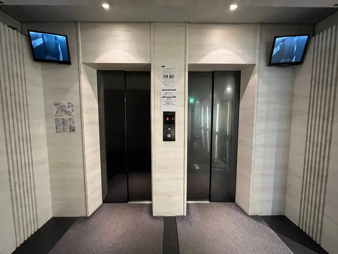 - モダンなエレベーターのロビー、テレビモニターが設置され、明るい照明の内装、壁には案内掲示物が貼られている