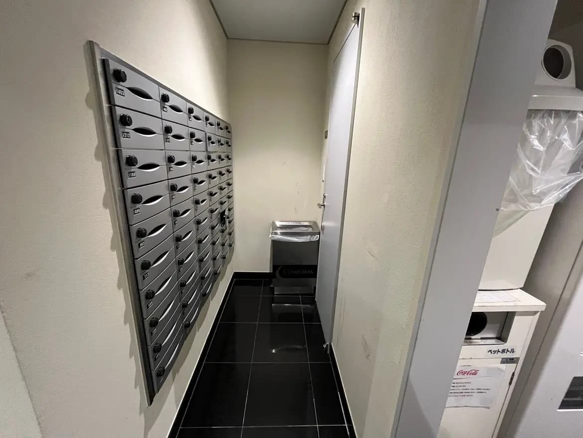 - 複数の郵便受けが並ぶ狭い廊下、奥にはゴミ捨て用の容器と白いドアがあるシンプルな空間
