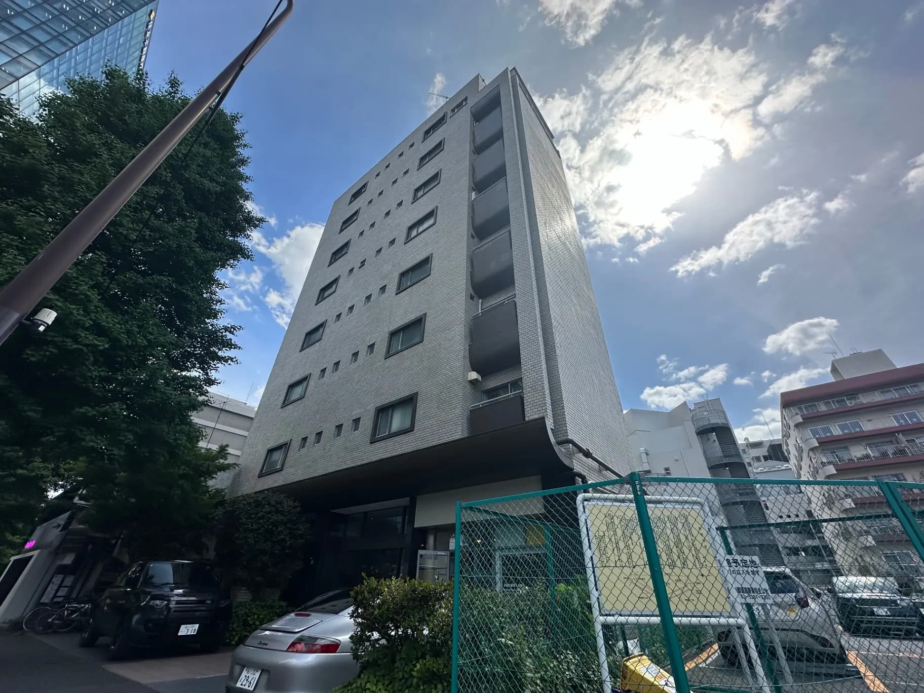 都心に位置するモダンなマンションビル、晴天の日差しと青空、周囲には緑と建物