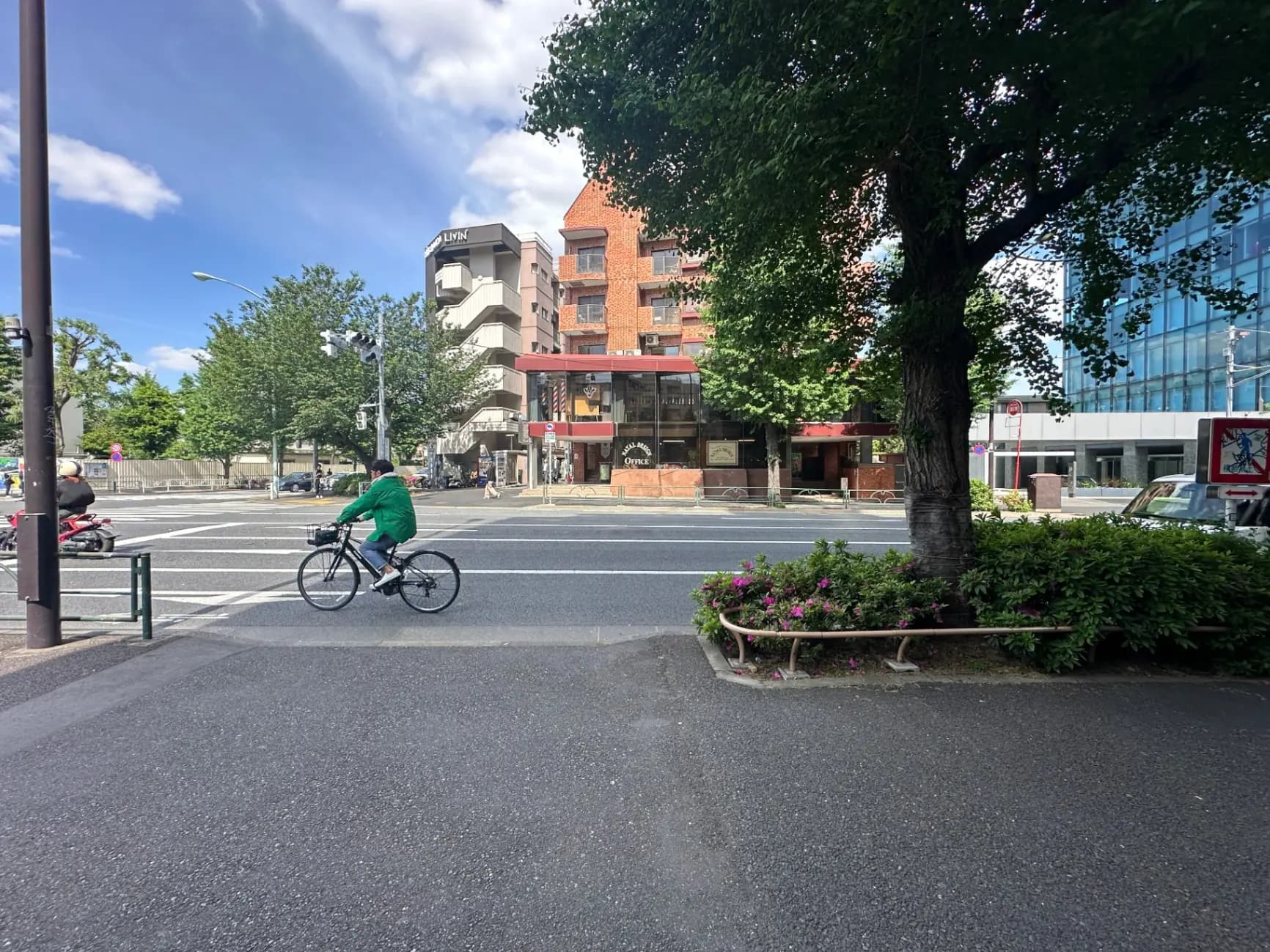 都市の交差点で自転車に乗る緑のジャケットの人、背景には赤レンガの建物とガラス張りのビル、花壇にはピンクの花が植えられている風景