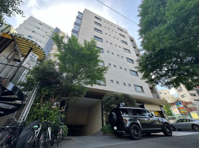 都会の景観に溶け込むモダンな建築物、駐車スペースに停められた高級SUV車、自転車が並ぶ歩道