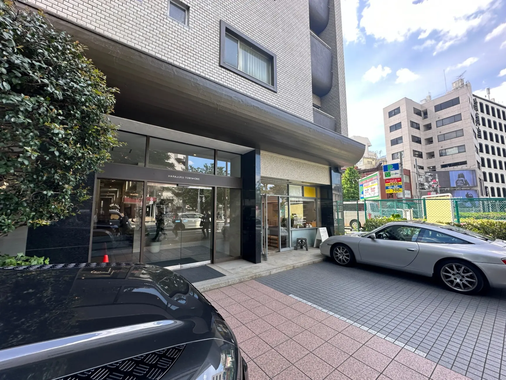 市街地にあるモダンな建物のエントランス、ショーウィンドウと駐車場、青空と高層ビルの風景