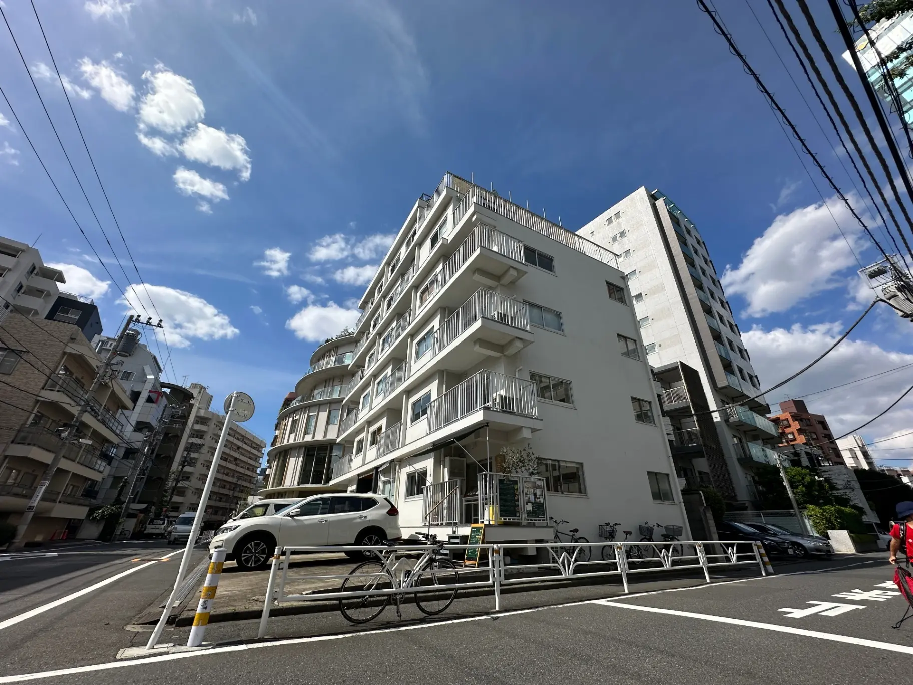 都会的な建物が建ち並ぶ街角、晴れた日の青空と白いマンション、街路樹と駐車中の車