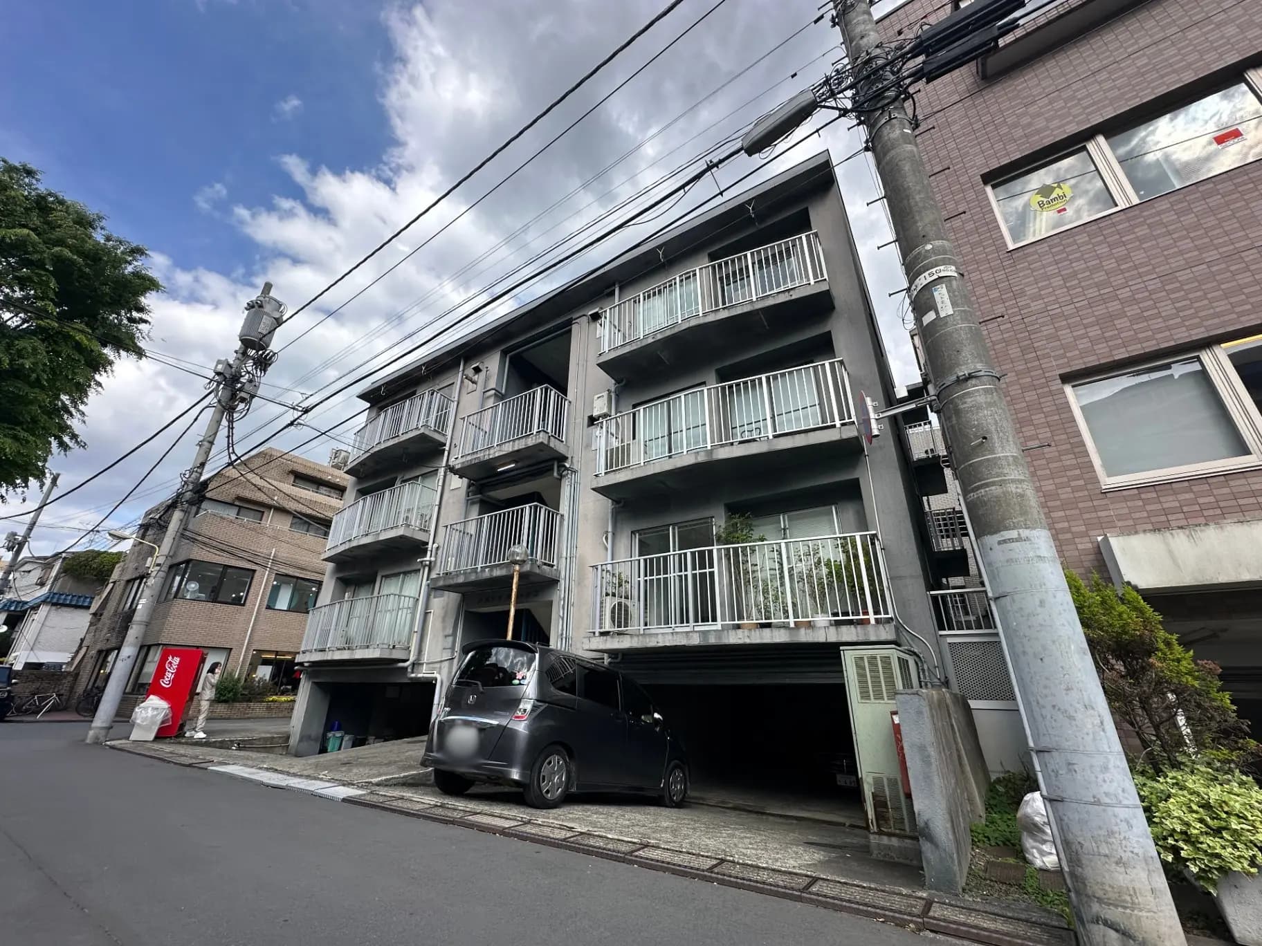 都市の住宅街にあるアパートメントビル、ベランダ付きの部屋と駐車スペース、通りには自動車と電柱