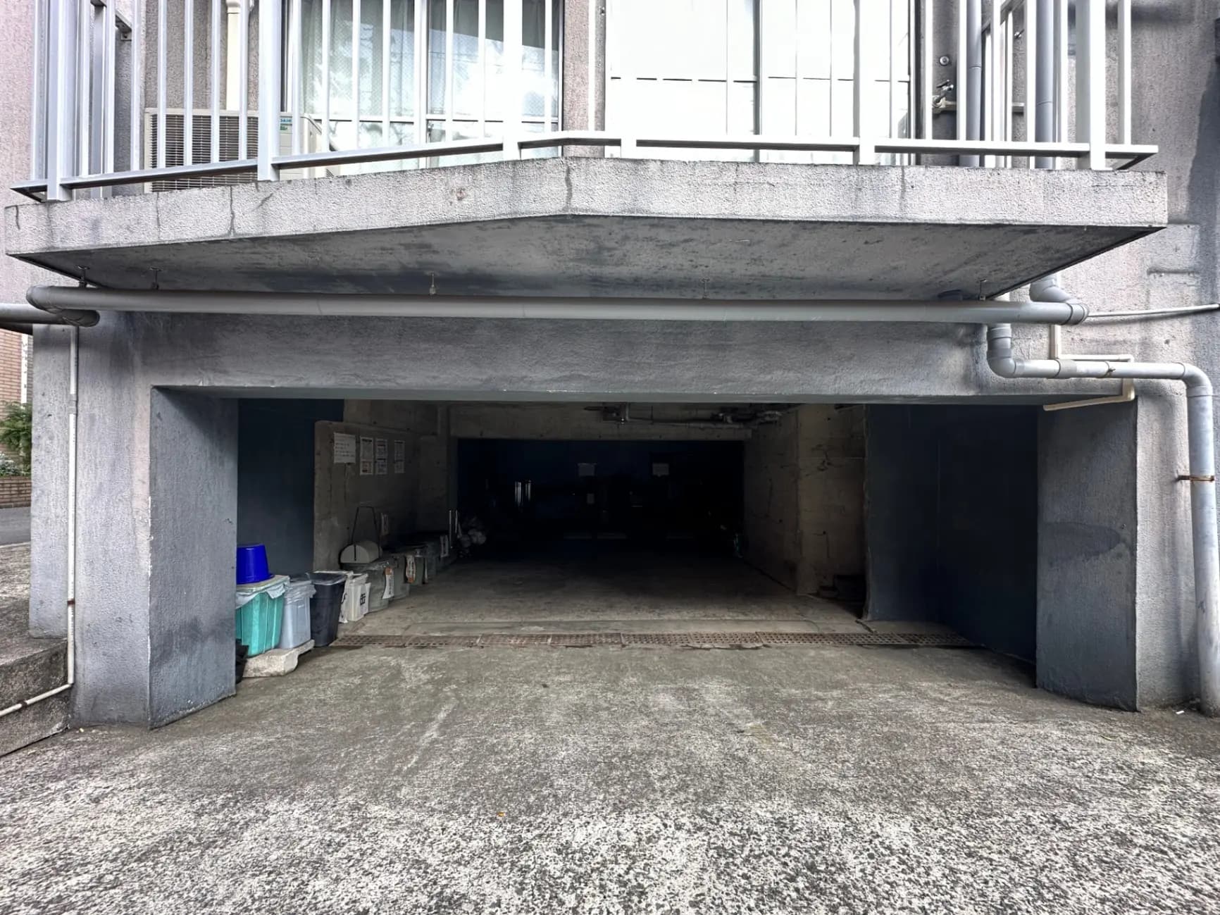 灰色のコンクリート製マンションの地下駐車場入り口、ゴミ箱と配管が見える