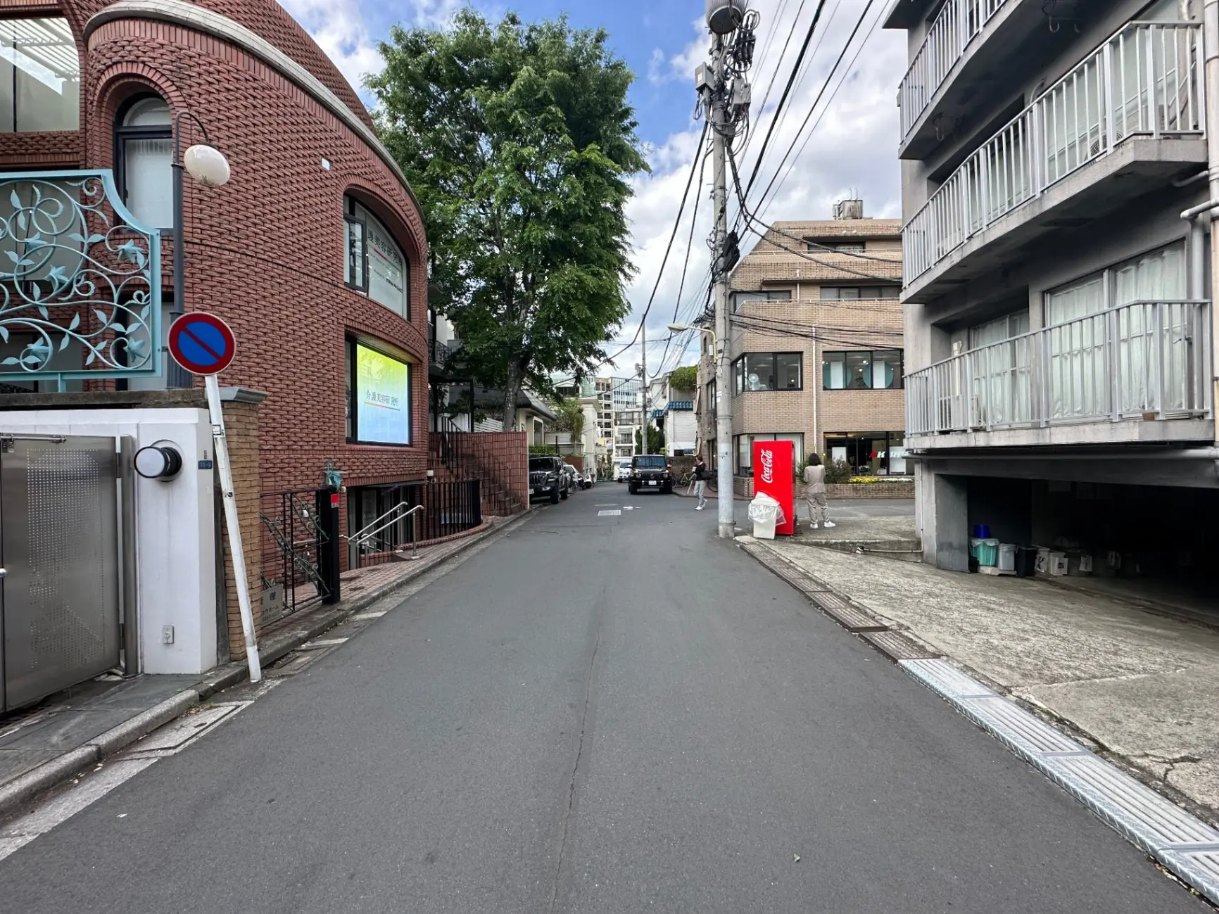 静かな住宅街の道路、レンガ造りの建物とアパート、街路樹と電柱、コカ・コーラの自動販売機が見える光景