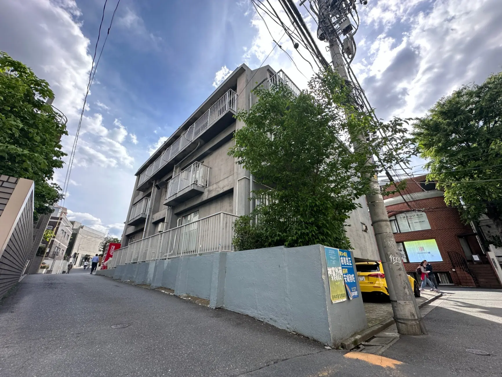 青空と雲間に覆われた都市の路地、コンクリートの質素な建物と電線、人々が歩く光景
