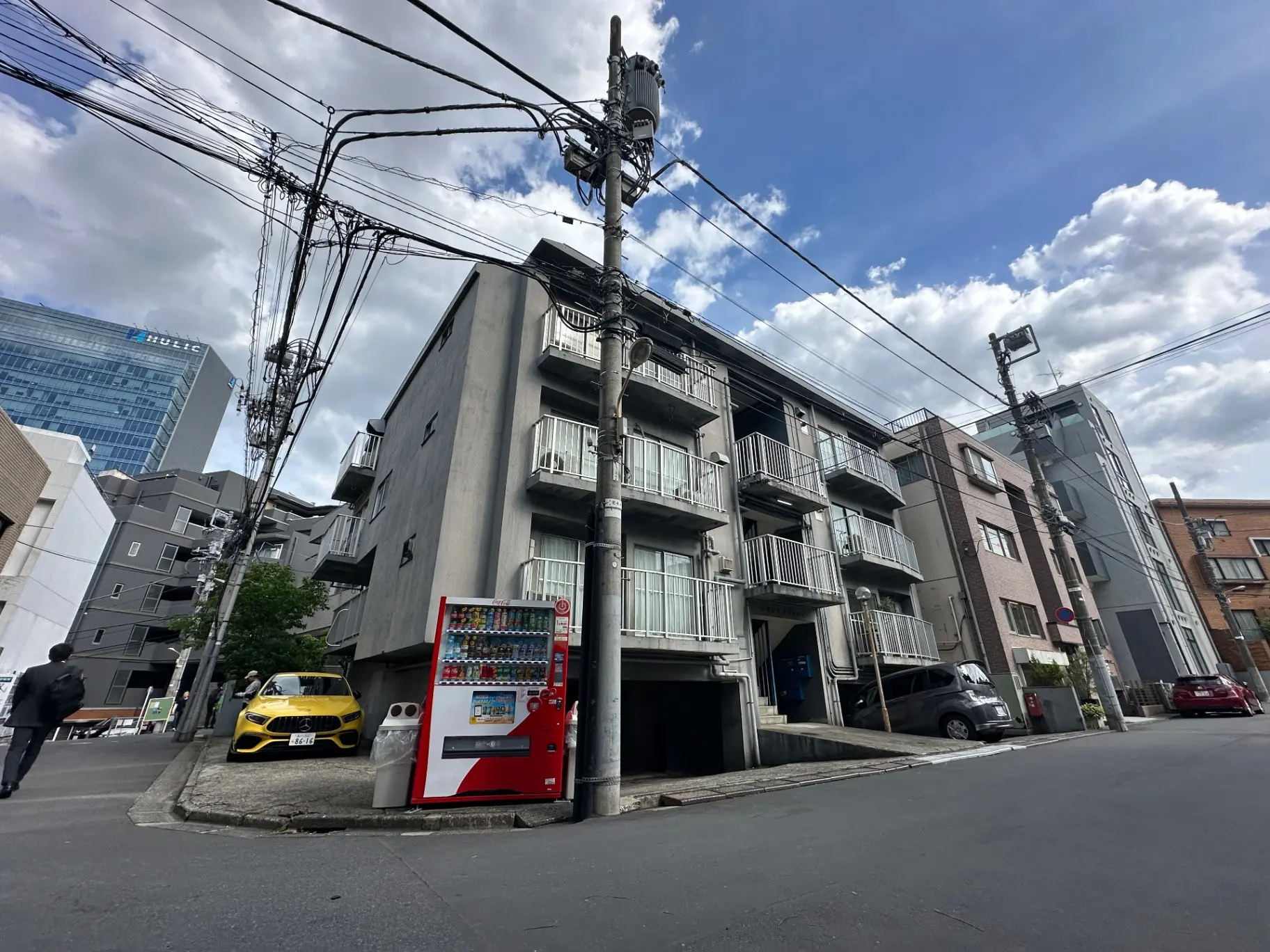 都市部のアパートの外観、駐車場に黄色いスポーツカー、背景に高層ビル、自動販売機が設置された歩道