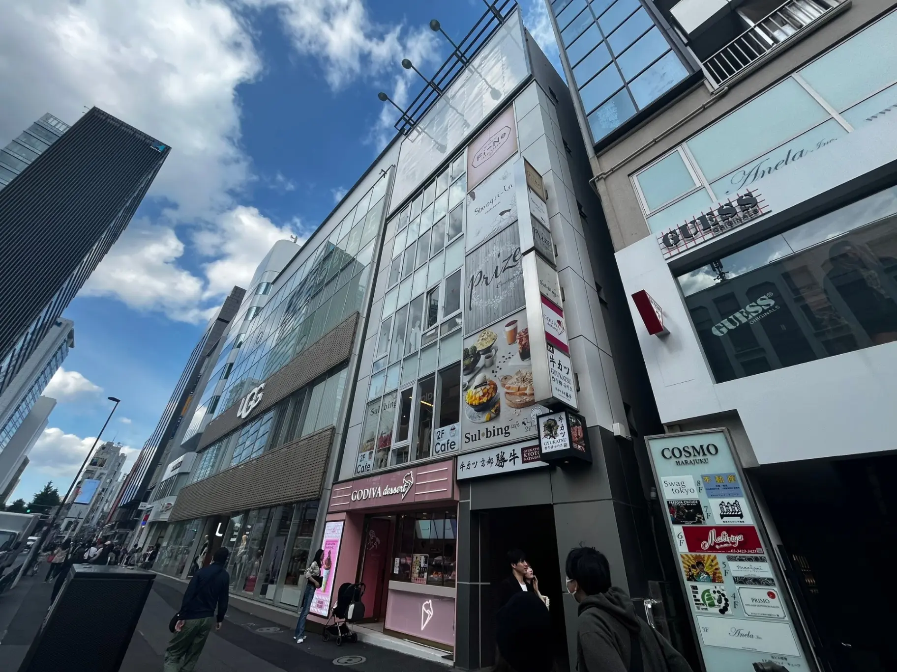 青空と雲が広がる都会の通り、多様なショップとレストランの看板が並ぶビルの外観