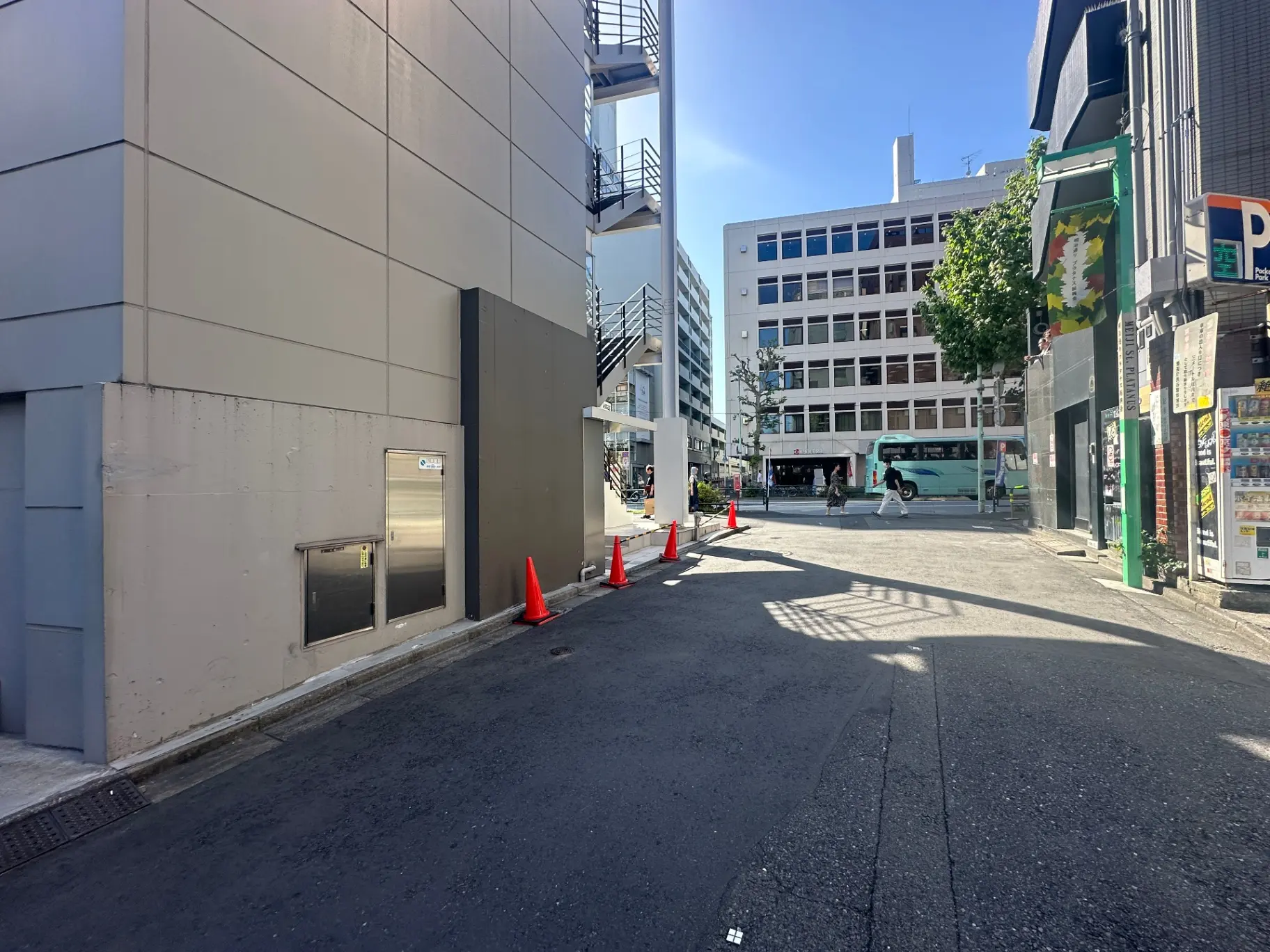 都会の路地裏、ビルの壁面、交通コーンが並べられている道路、遠くにバスが停車している風景