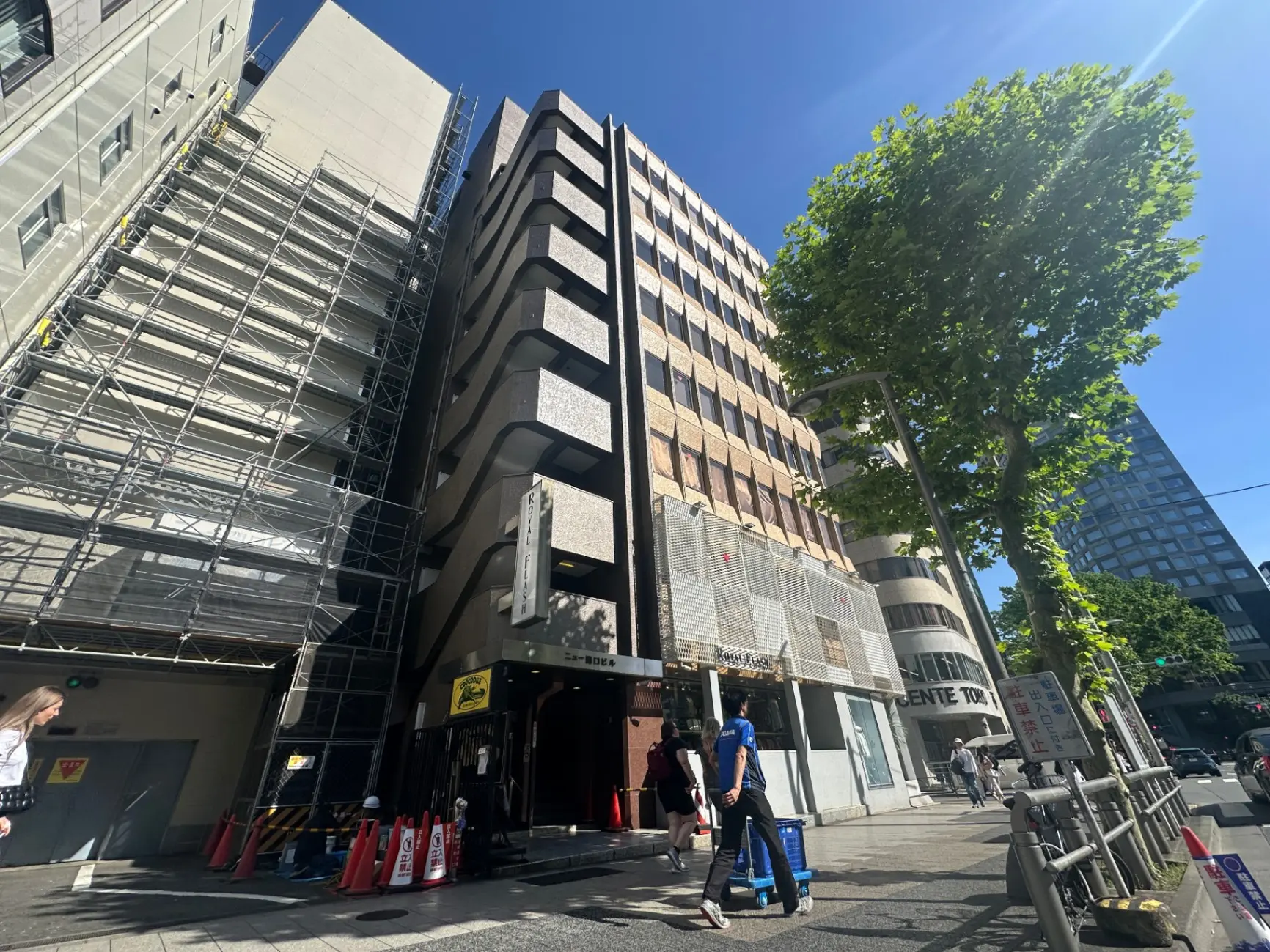 日差しが照りつける都会の街角、足場が組まれたオフィスビルの外観と歩行者