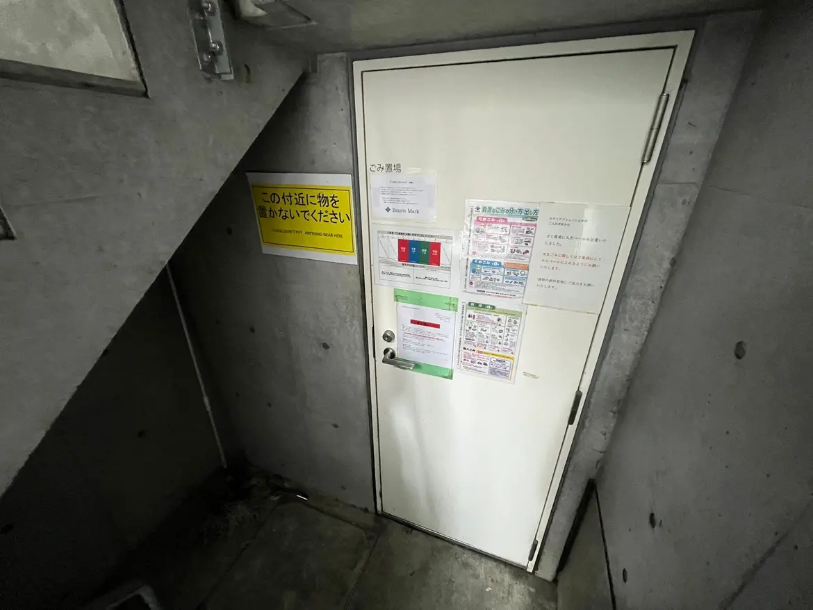 - コンクリートの壁に囲まれた階段の途中にある白いドア、周囲に掲示物と注意書きが貼られている風景