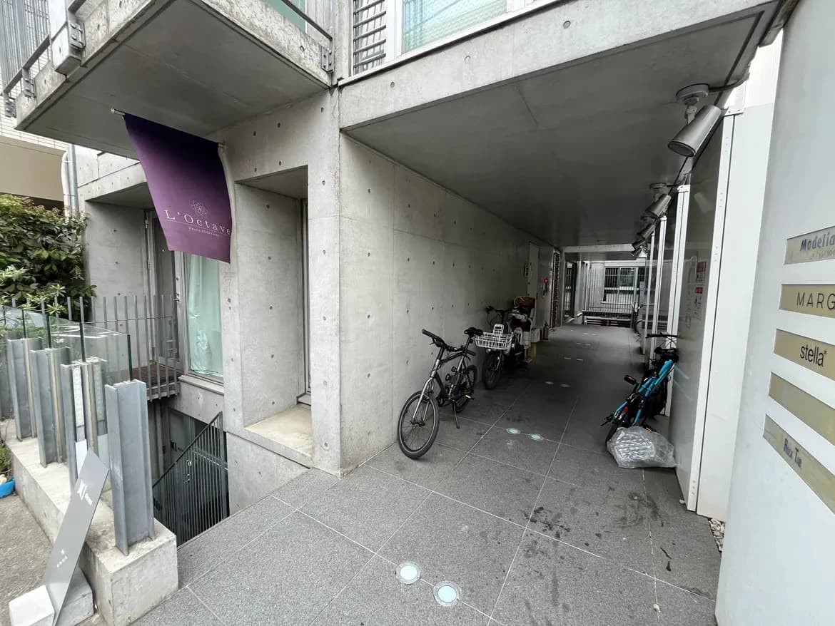 - コンクリートの壁とグレーのタイルの通路、左側に店舗の看板「L'Octave」、自転車と荷物が置かれたエリアの外観