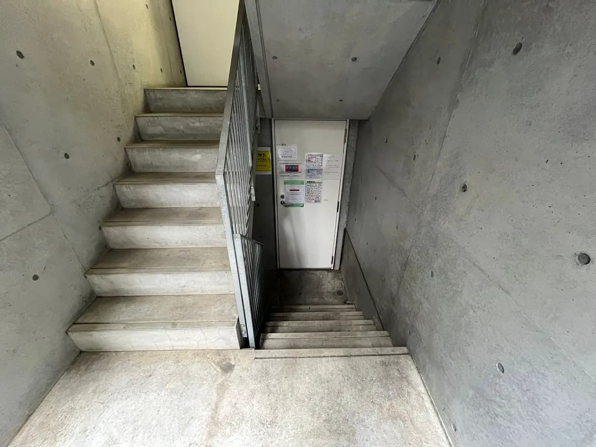 - コンクリートの壁と階段が特徴的なシンプルなエントランス、扉には掲示物が貼られている様子