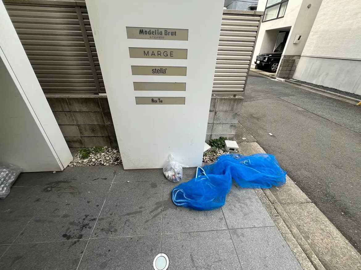 - モデルブリュットのエントランスサイン、散らばった青いゴミ袋と周囲の舗装された歩道が映る街の風景