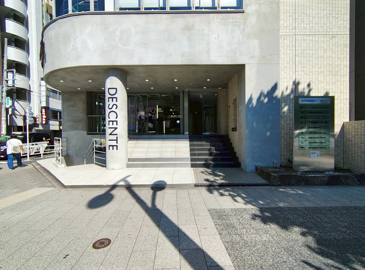 - モダンな外観のデサント店舗入り口、大きな柱に「DESCENTE」のロゴ、階段と周辺の街並みが見える風景