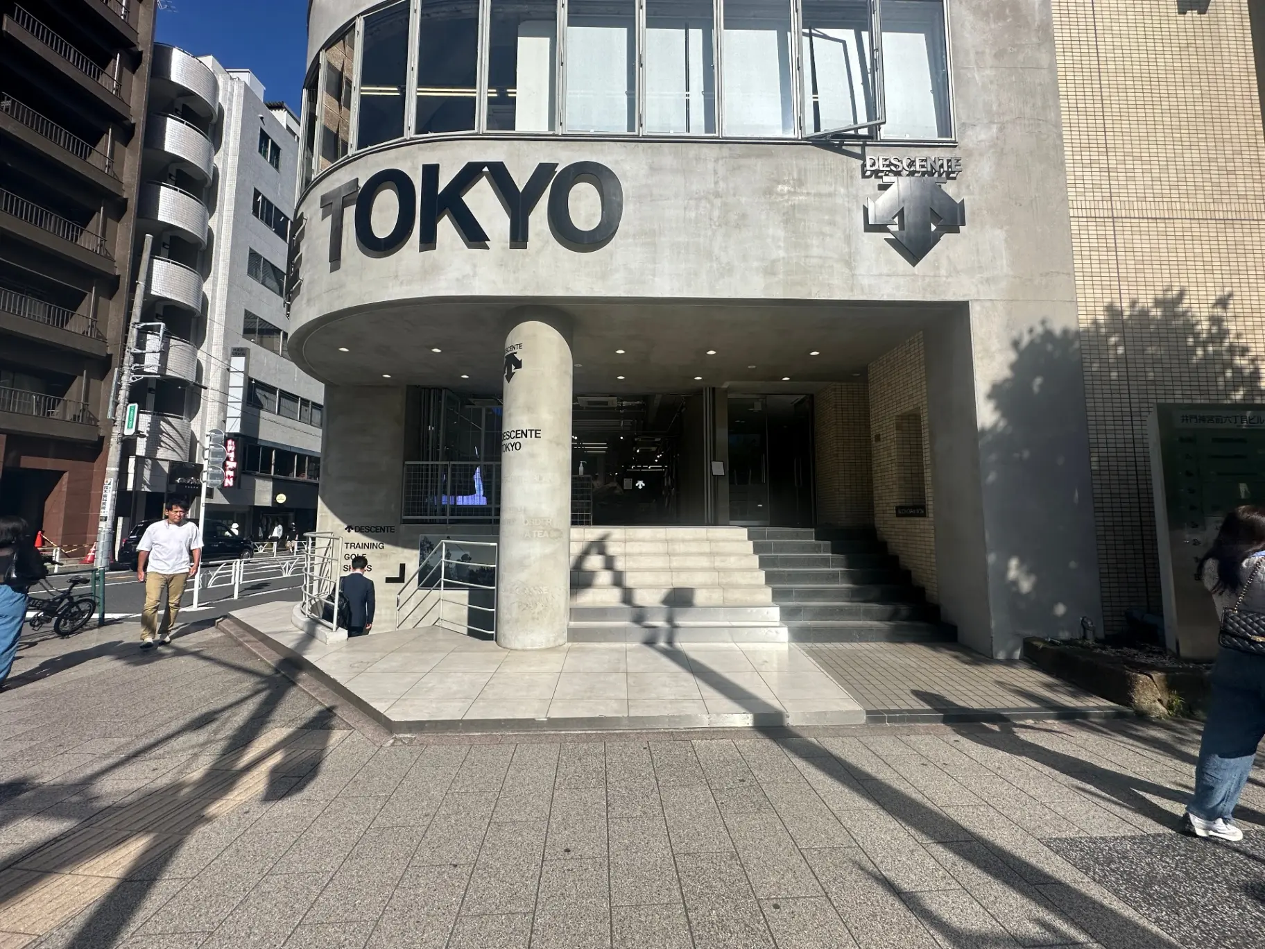日光が差し込む晴れた日の東京のビル街、歩行者と自転車が通る歩道、目立つ大きなTOKYOのサインがある建物の入り口