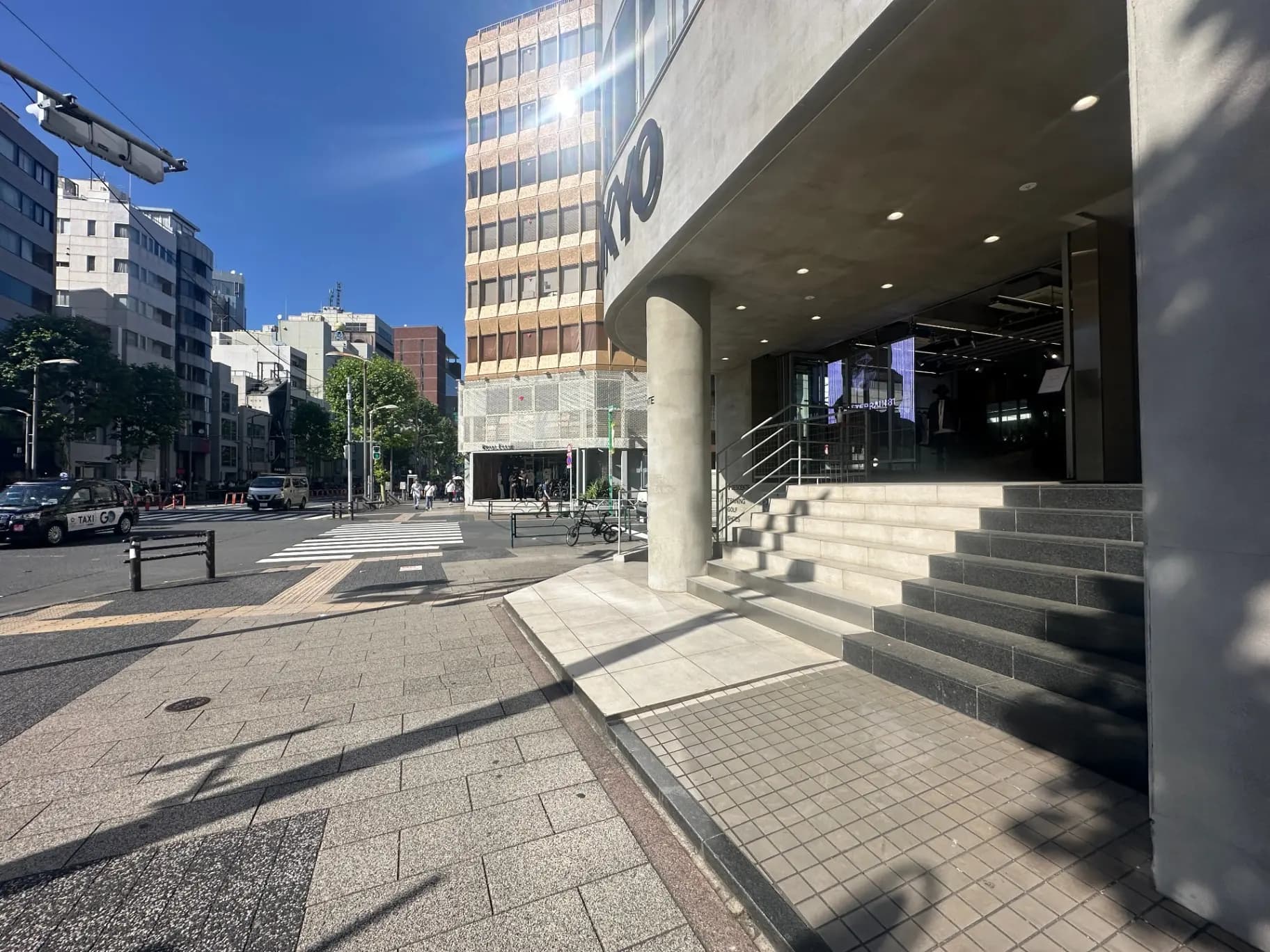 都市の交差点に面したモダンなビルのエントランス、日差しの中で歩行者が通り過ぎる光景