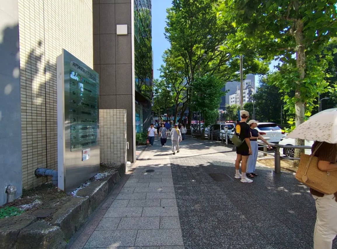 - 日差しが降り注ぐ都市の歩道、木々に囲まれた街並みで人々が行き交う風景、近くのビルの案内板が見える