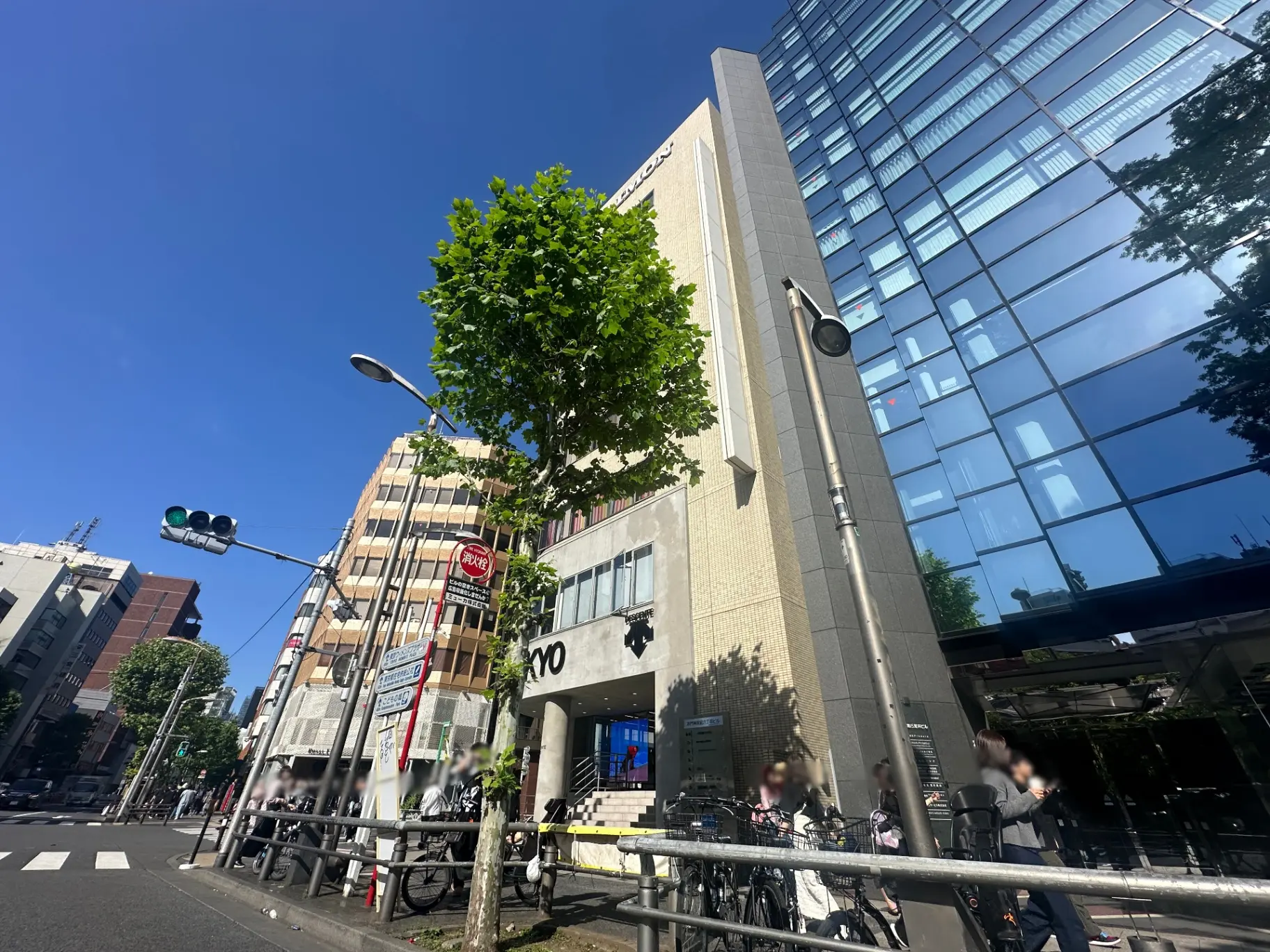 都会的な通りの風景、青空と緑の木々、現代的なビルと商業施設、歩行者と自転車が行き交う光景