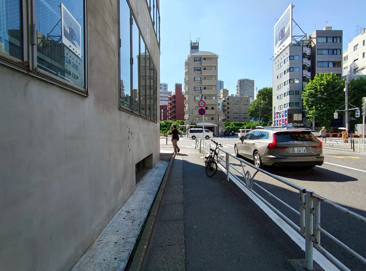 - 都会の街並み、ビルの隙間にある歩道、自転車が停められた側道と通行中の車両の風景
