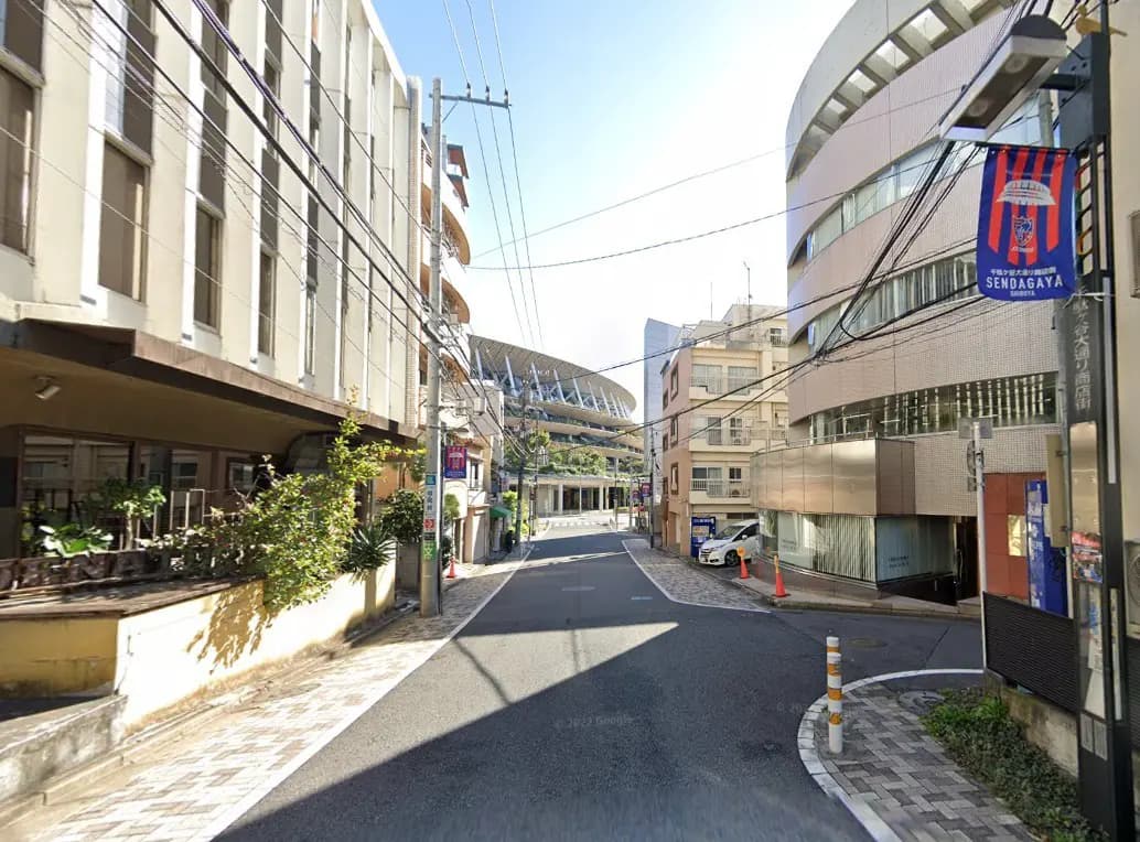 - 送信ヶ谷の街並み、道路に沿った商業ビルと住宅、遠くに体育館の屋根が見える風景