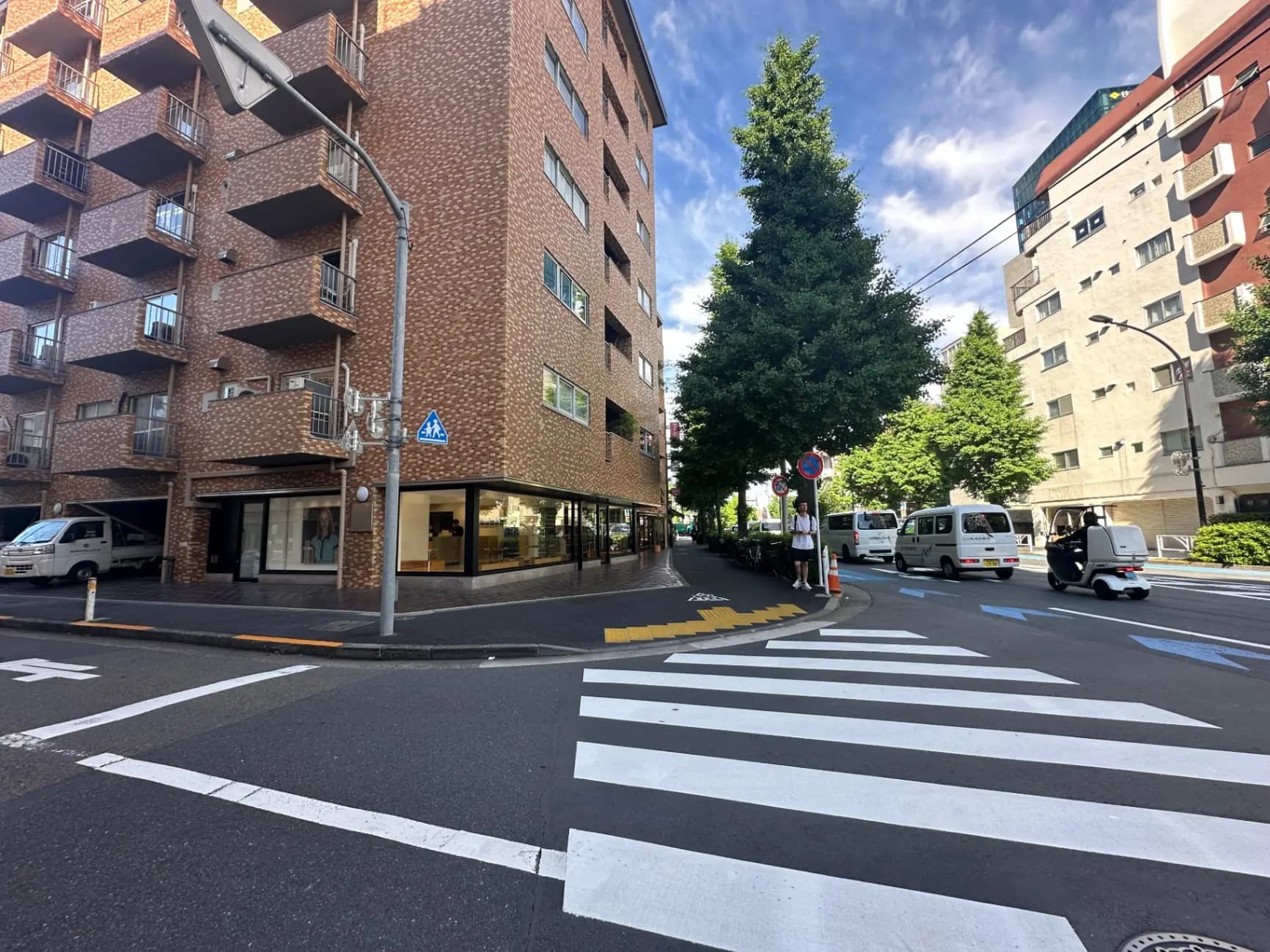 青空の下、都市の交差点と横断歩道、茶色いレンガ造りの建物と緑の木々が並ぶ通り