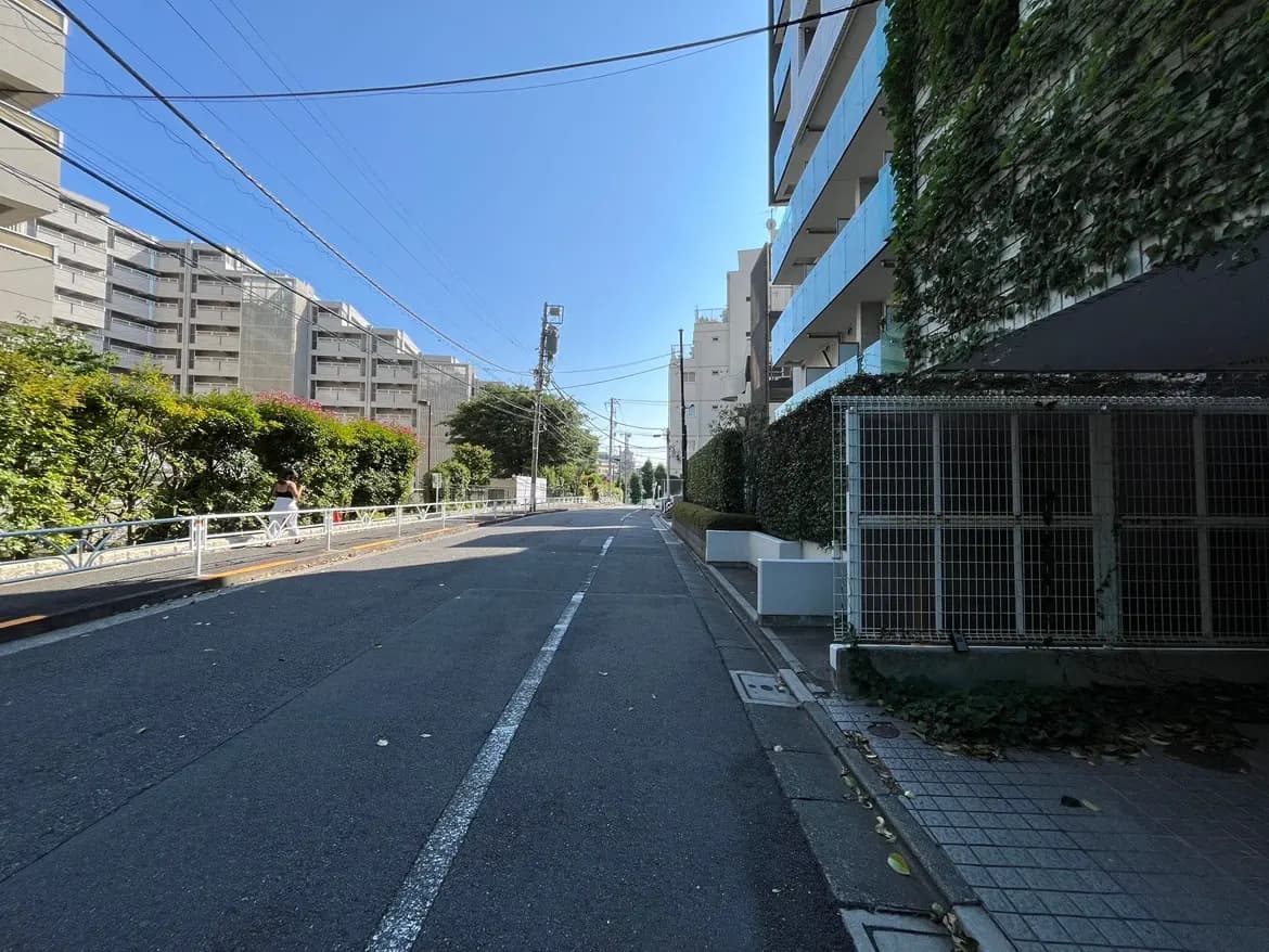 - 明るい青空の下、整備された道路沿いに並ぶマンションと緑豊かな植栽、歩行者が通る様子が映った街の風景