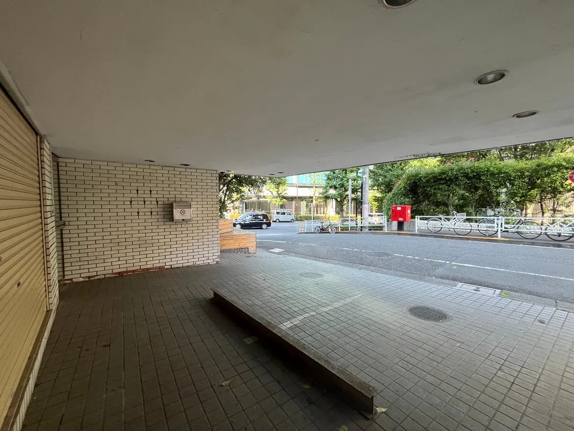 - 街に面したシャッターの閉じた店舗、側にベンチと郵便ポスト、周囲の緑が目を引くエントランスエリア