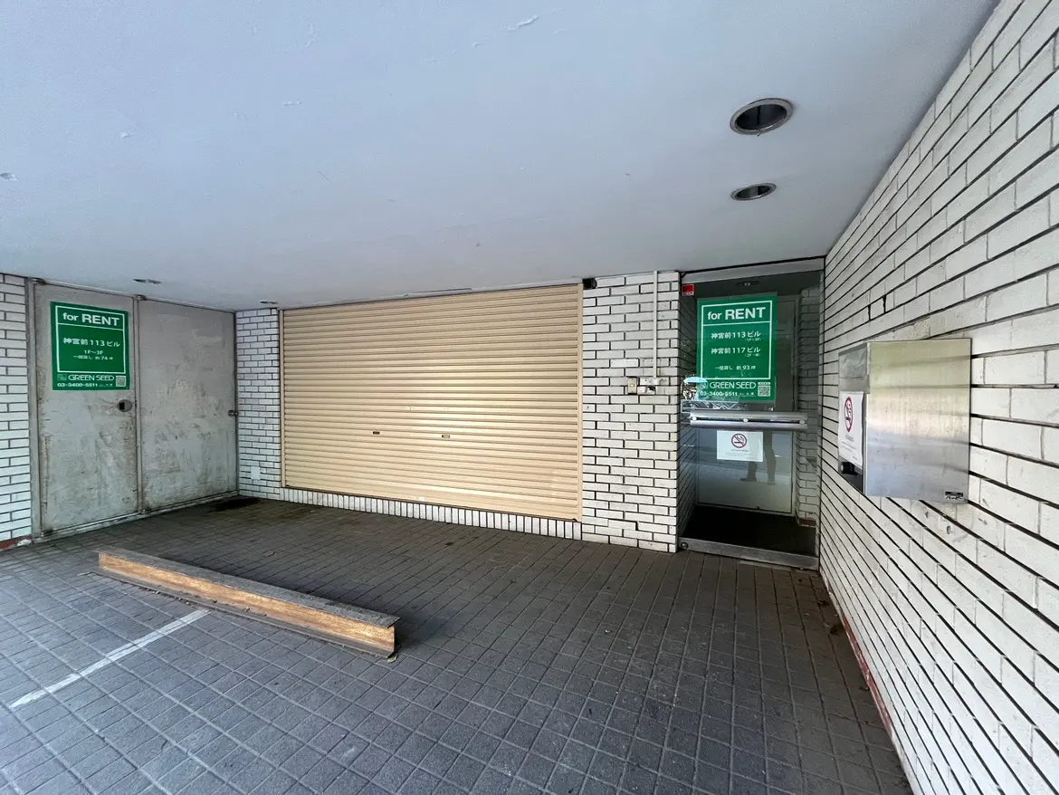 - 賃貸物件のエントランス、ブロックタイルの床、閉まったシャッターと賃貸広告の看板がある商業スペース