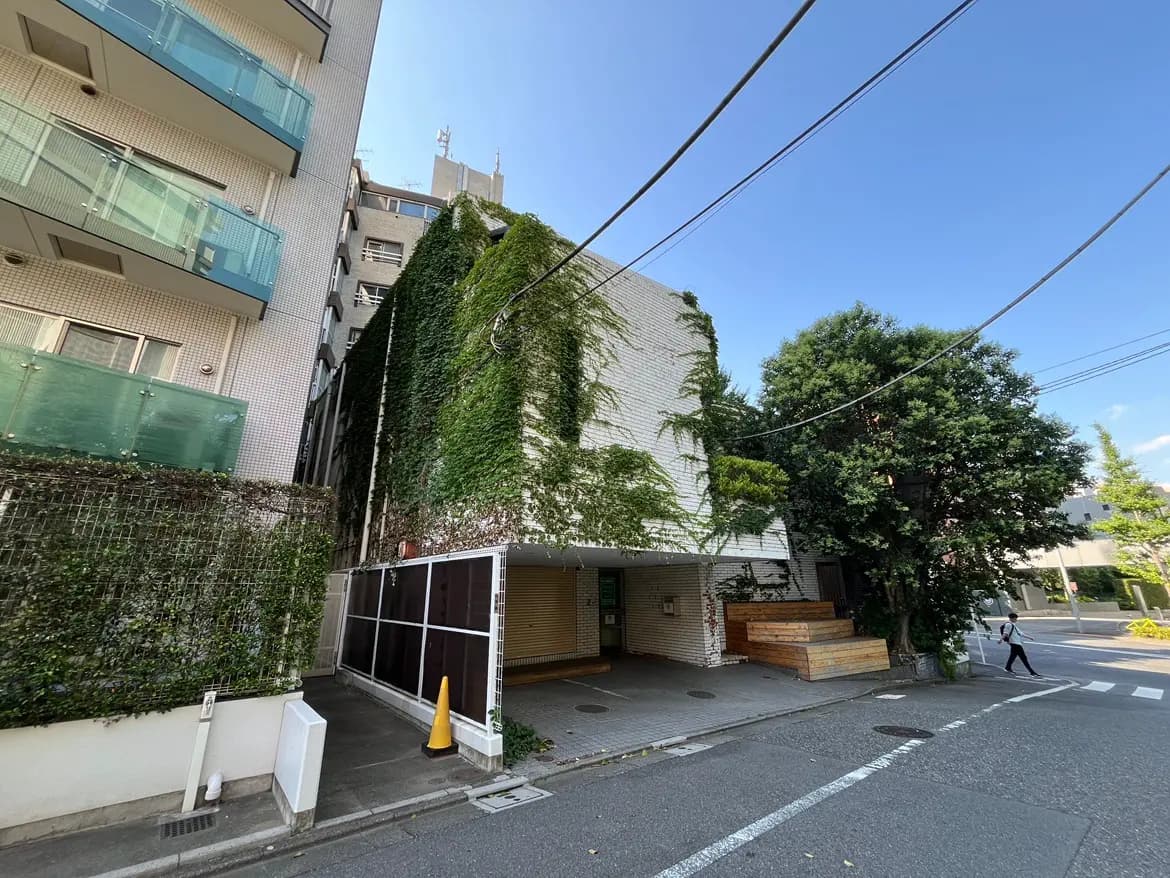 - 緑のつる植物が生い茂る建物の外観、周囲に木々があり、青空が広がる住宅街の風景