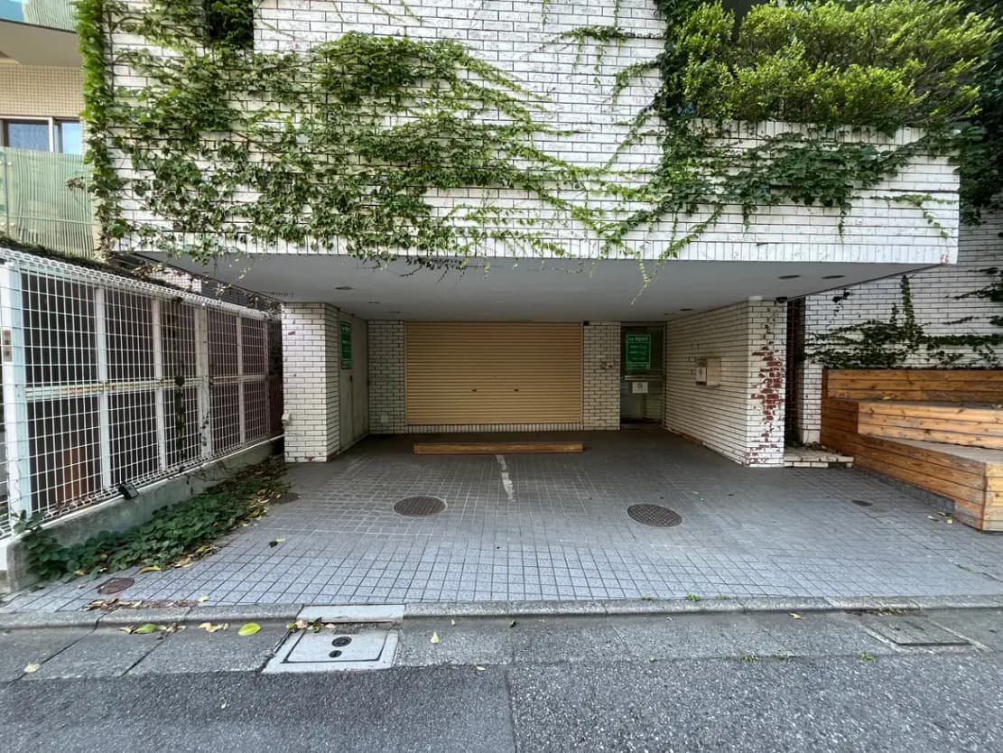 - 緑豊かな植物が絡まる白レンガの壁面と、シャッター付きの駐車場、周囲に広がる平坦な舗装のエリア