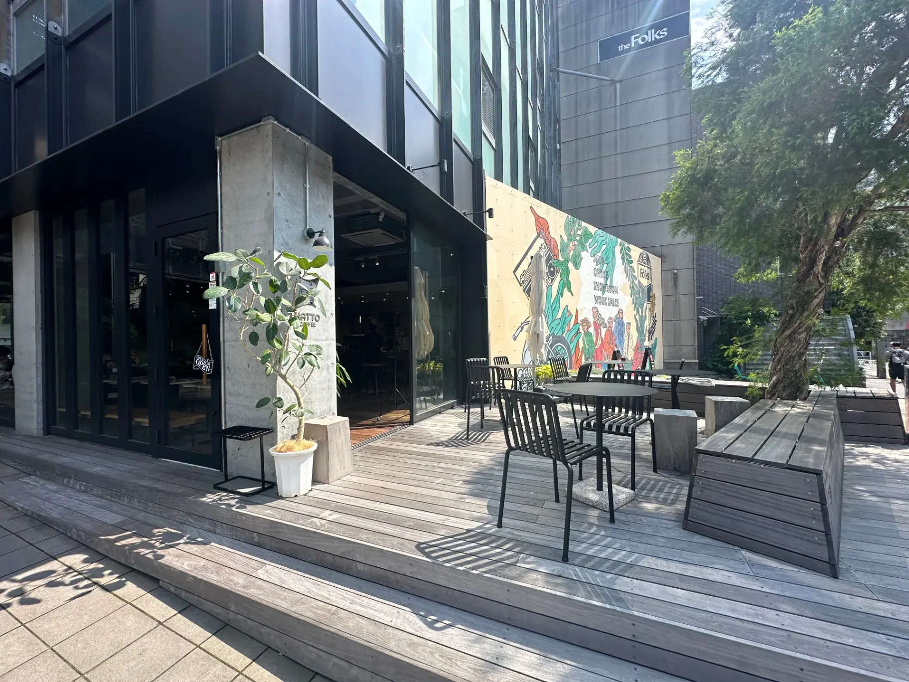 モダンなカフェの屋外席、木製デッキとテーブル、壁画アート、日差しが降り注ぐ快適な休憩スペース