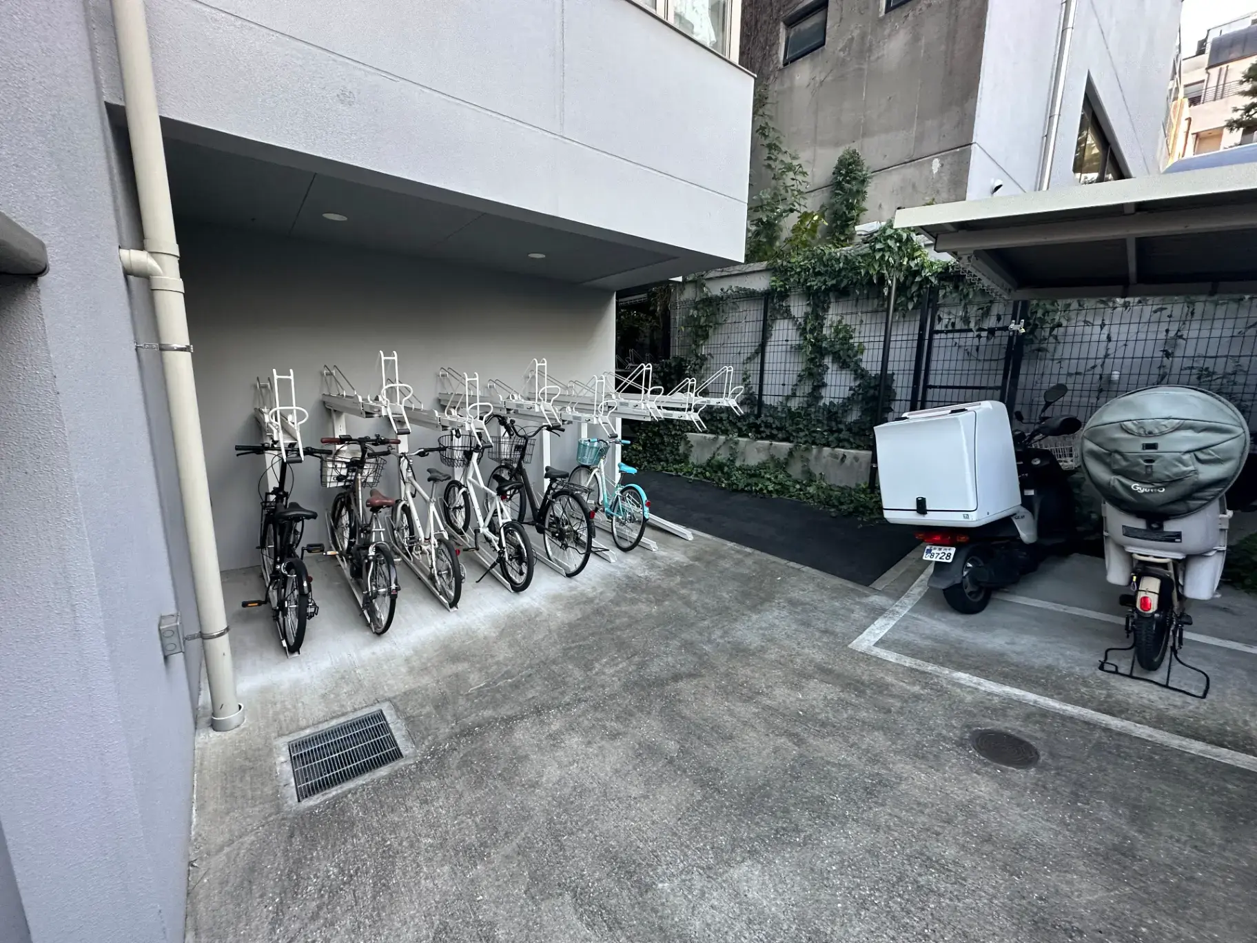 建物の軒下に設置された自転車駐輪スペース、整理された自転車と配達用スクーター、緑の植物がある都市の屋外パーキングエリア
