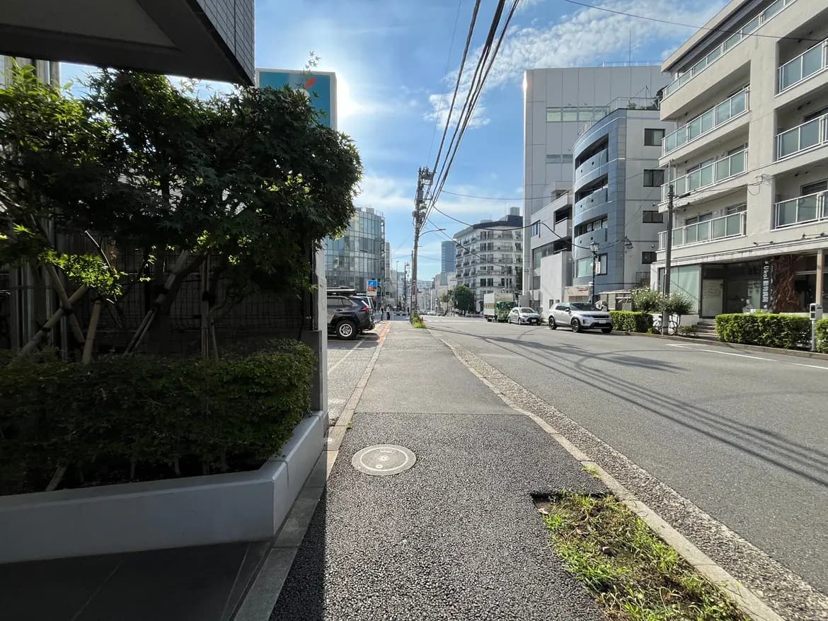- 都市の街並みを映した道路の風景、青空の下に続く歩道と車の並ぶ様子、周囲の建物と緑の植栽が特徴的な風景