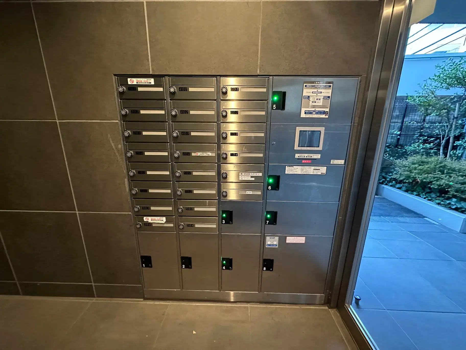 マンションの共用エントランスホールに設置された銀色の郵便受け、番号ラベルとセキュリティカメラ付き