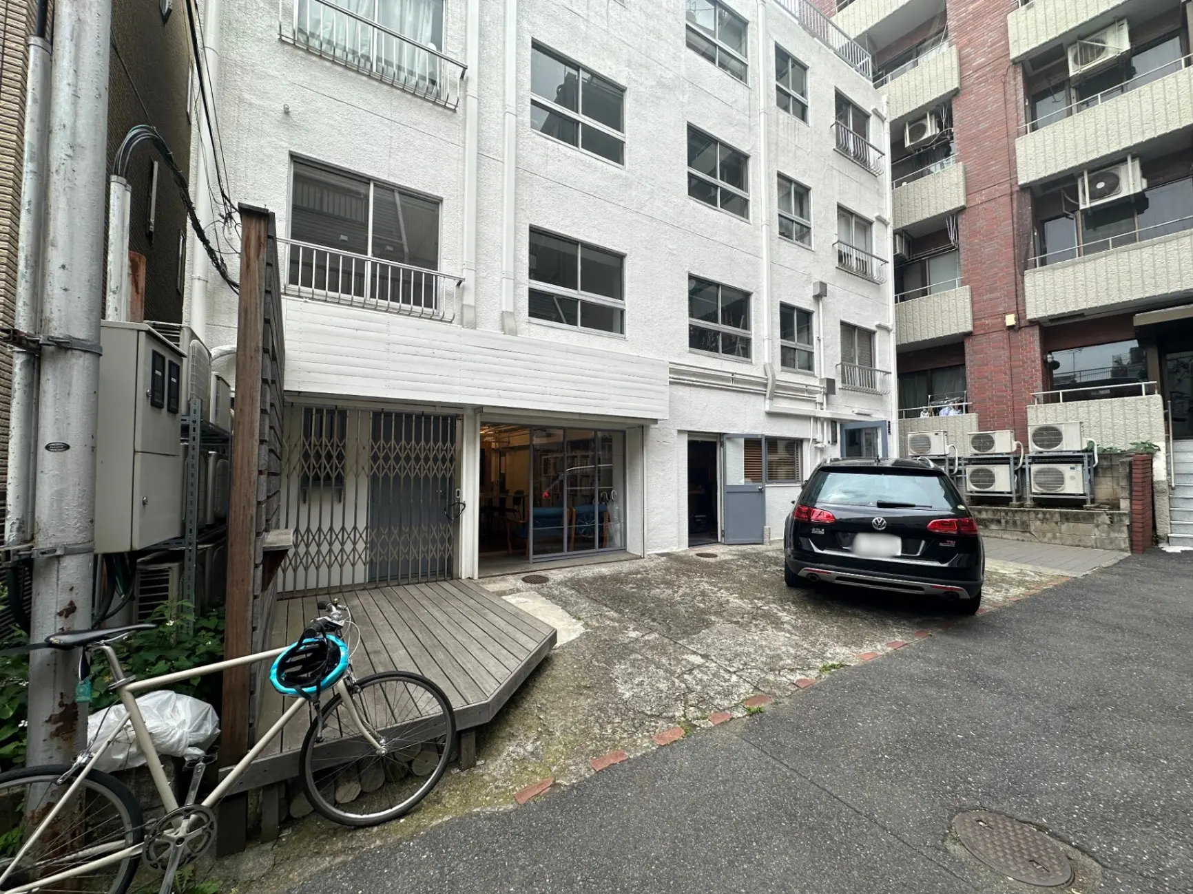 都市のアパートの駐車場、自転車と黒い車が停まっている、裏口とエアコンの外部ユニットが見える