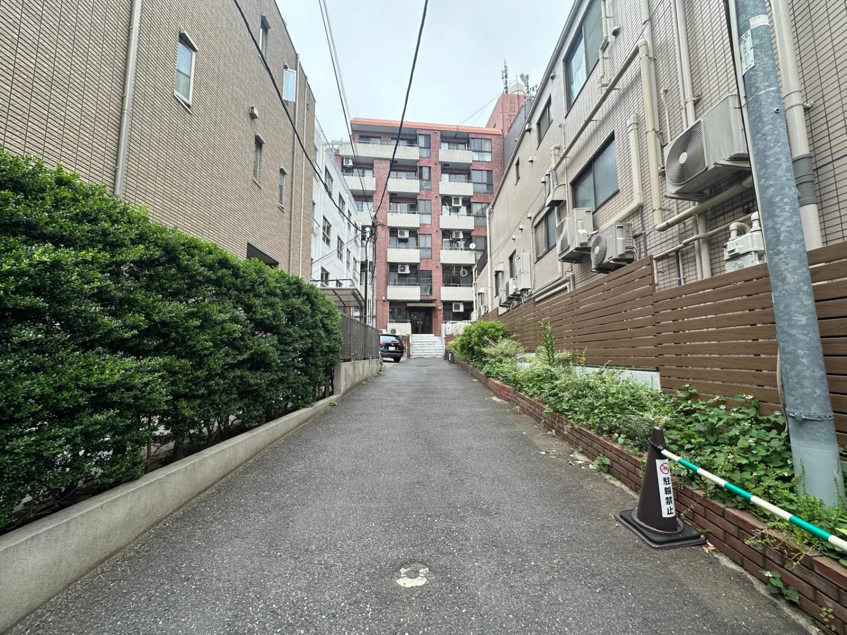 都市のアパートメント脇の細い舗装された道、緑の植え込みと複数のエアコン外部機器が見える景観