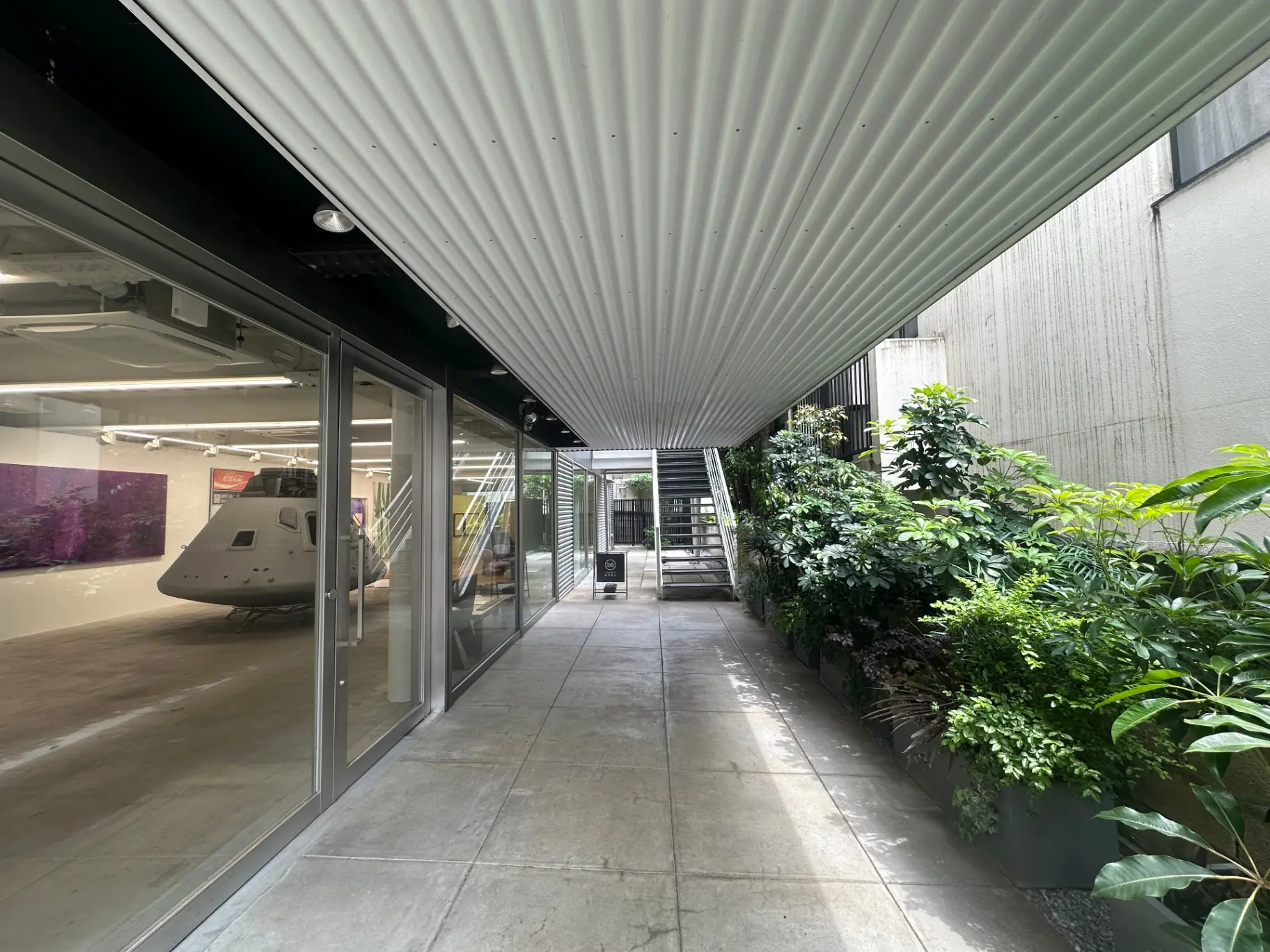 モダンなオフィスビルの屋外パティオ、天井は波型の金属製、壁には緑豊かな植物、ガラス扉と宇宙船の模型が見えるデザイン