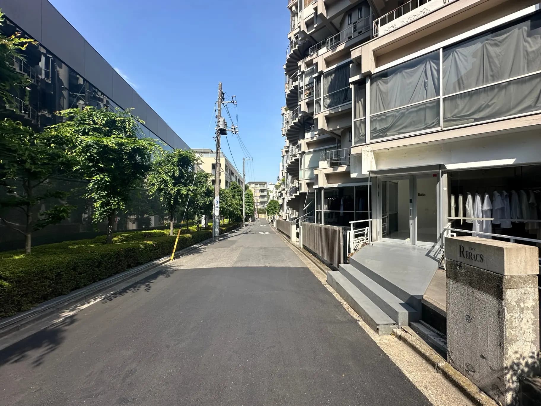 晴れた日の住宅街の道、左側には緑豊かな植栽とモダンな外観の建物、右側にはマンションの入り口と並木道