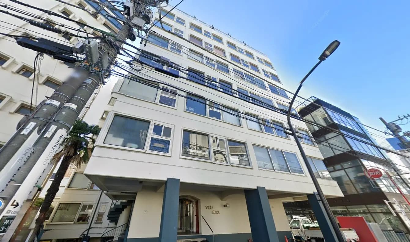 都市の街並みに建つ多階建てのモダンなアパート、青空とパームツリー、周囲には電柱と電線が見える景観