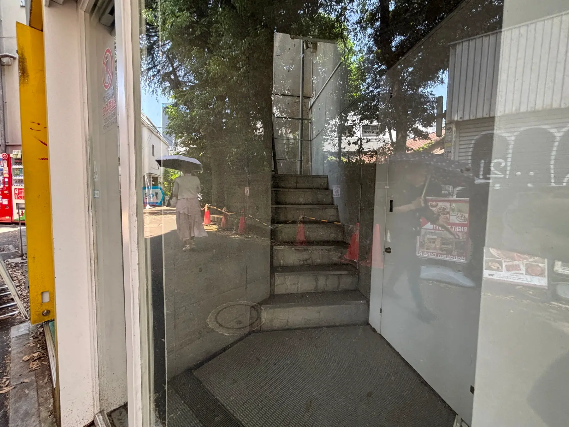 ガラス張りの建物の入口、階段が映り込んでおり、外を傘を持った人が歩いている光景を捉えた画像