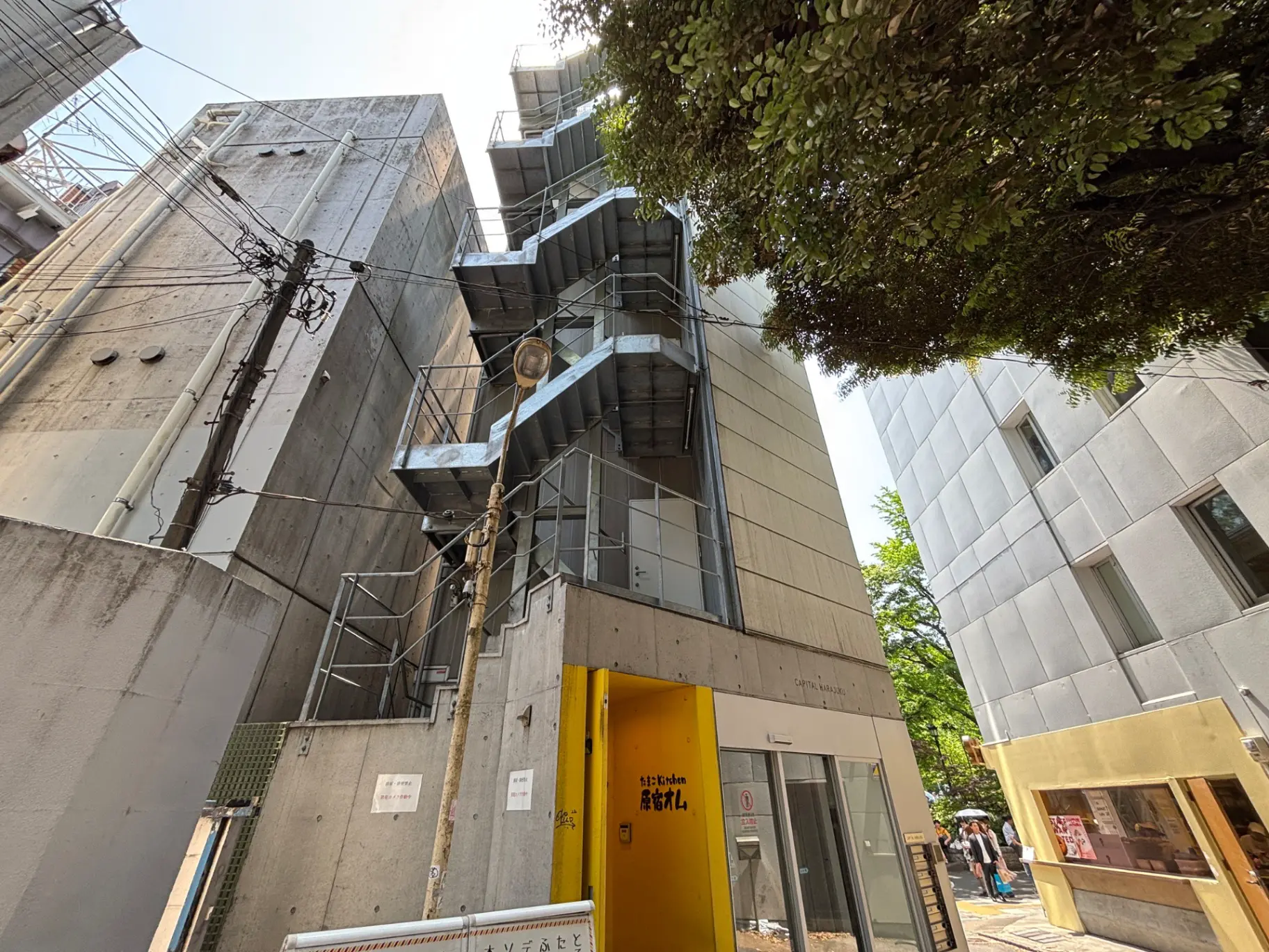 - コンクリート製の現代的な建物の外観、階段の手すりと明るい黄色の出入口、周囲に緑が見える都市風景